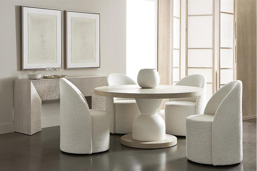 Four curved white upholstered chairs around a circular table with a white central base on a circular stone piece. The tabletop is a brown circular top and a decorative white vase infront of a stone console and two cream and white paintings